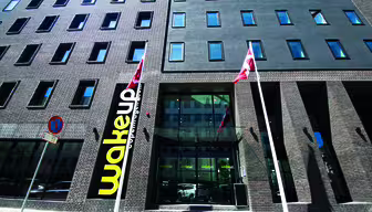 Gadebillede af en moderne murstensbygning med store vinduer. To flag er foran, og på et skilt står "vågning" ved indgangen. Et "parkeringsforbud"-skilt er også synligt på fortovet.