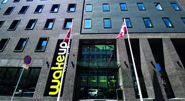 Gadebillede af en moderne murstensbygning med store vinduer. To flag er foran, og på et skilt står "vågning" ved indgangen. Et "parkeringsforbud"-skilt er også synligt på fortovet.