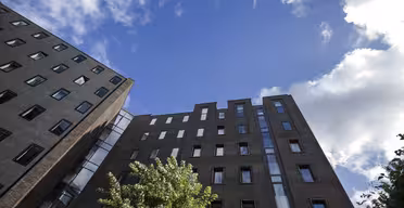 Opadgående udsigt over to høje bygninger med mørke murstens-ydre og flere vinduer, sat mod en lys blå himmel med spredte skyer. Et træ med grønt løv er synligt i forgrunden.
