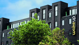 Moderne sort bygning med gult "Wakeup Copenhagen"-logo, med flere vinduer sat mod en blå himmel. Grønne træer i forgrunden tilføjer kontrast og tekstur til scenen.