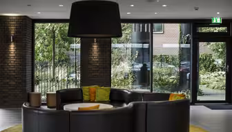 Moderne lobby med cirkulære siddepladser i sort læder omkring et hvidt bord, fremhævet af grønne og orange puder. En stor sort pendel hænger over hovedet. Gulv-til-loft vinduer giver udsigt til frodige grønne områder udenfor.