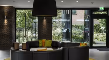Moderne lobby med cirkulære siddepladser i sort læder omkring et hvidt bord, fremhævet af grønne og orange puder. En stor sort pendel hænger over hovedet. Gulv-til-loft vinduer giver udsigt til frodige grønne områder udenfor.