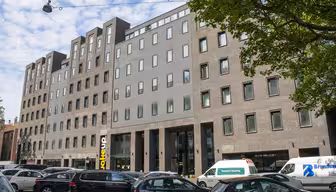En moderne etagebyggeri med en blanding af grå og brun murstensfacade. Store vinduer kanter strukturen. Biler er parkeret foran, og træer indrammer delvis scenen. En blå himmel med spredte skyer er synlig ovenfor.