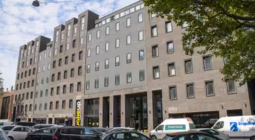 En moderne etagebyggeri med en blanding af grå og brun murstensfacade. Store vinduer kanter strukturen. Biler er parkeret foran, og træer indrammer delvis scenen. En blå himmel med spredte skyer er synlig ovenfor.