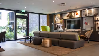 Moderne loungeområde med en lang grå sofa med grønne og brune stole. Værelset har et stort TV, dekorative reoler og udsigt til en frodig have gennem glasdøre. Gulvet er flisebelagt med et brunt tæppe under siddeområdet.