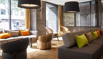 Moderne lobby med grå sofaer prydet med grønne og orange puder. Brune læderstole omkranser små borde. Store sorte pendellamper hænger fra loftet. Naturligt lys strømmer ind gennem store vinduer og oplyser rummet.