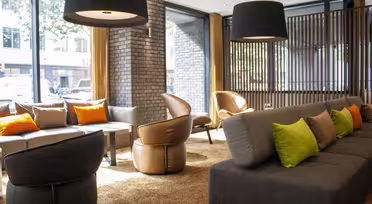 Moderne lobby med grå sofaer prydet med grønne og orange puder. Brune læderstole omkranser små borde. Store sorte pendellamper hænger fra loftet. Naturligt lys strømmer ind gennem store vinduer og oplyser rummet.
