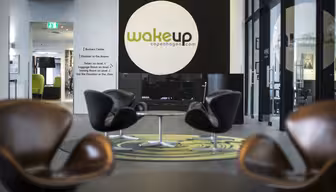 Moderne hotelobby med slanke brune stole omkring runde borde. På et stort rundt skilt står der "wakeup copenhagen.com" på en mørk væg. Lyst, luftigt rum med gulv til loft-vinduer og et grønt tæppedesign.