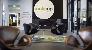Moderne hotelobby med slanke brune stole omkring runde borde. På et stort rundt skilt står der "wakeup copenhagen.com" på en mørk væg. Lyst, luftigt rum med gulv til loft-vinduer og et grønt tæppedesign.