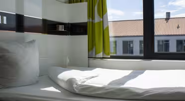 Minimalistisk værelse med en enkeltseng dækket af hvide lagner og en pude ved siden af et stort vindue med grønne og hvide gardiner. Sollys strømmer ind og oplyser rummet. Uden for vinduet ses bygninger med røde tage.