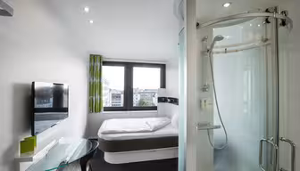 Et moderne hotelværelse med et kompakt design med en seng mod væggen, et vægmonteret tv, et skrivebord med glasplade med en stol og en buet glasindkapslet bruser. To store vinduer giver udsigt til bygninger udenfor.