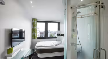 Et moderne hotelværelse med et kompakt design med en seng mod væggen, et vægmonteret tv, et skrivebord med glasplade med en stol og en buet glasindkapslet bruser. To store vinduer giver udsigt til bygninger udenfor.