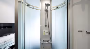 En moderne, glasindlukket bruser med buede, frostede døre. Indvendigt er der et slankt brusehoved og kontrolpanel. Det udvendige område viser en del af et værelse med sort/hvid farveskema.