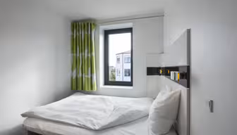 Et lille, minimalistisk soveværelse med en hvid seng, pænt hvidt sengetøj og to puder. Et vindue med grønne og hvide gardiner viser udsigt til nabobygninger under en klar himmel. Hvide vægge forstærker rummets lysstyrke.