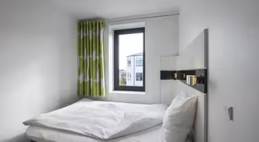 Et lille, minimalistisk soveværelse med en hvid seng, pænt hvidt sengetøj og to puder. Et vindue med grønne og hvide gardiner viser udsigt til nabobygninger under en klar himmel. Hvide vægge forstærker rummets lysstyrke.