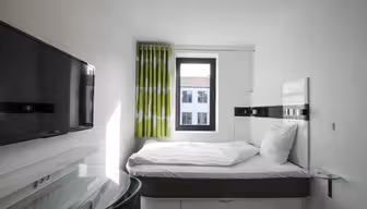 Lille moderne hotelværelse med en seng ved et vindue, med grønmønstrede gardiner og udsigt til nabobygningerne. Et vægmonteret tv er synligt til venstre, og et skrivebord med glasplade er i forgrunden.