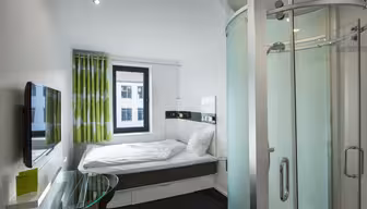 Kompakt moderne hotelværelse med en enkeltseng, hvidt sengetøj og gardiner med grønne accent. Der er et vægmonteret tv, et glas skrivebord og en buet glas brusekabine til højre. Et vindue har udsigt over en bygnings ydre.
