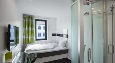 Kompakt moderne hotelværelse med en enkeltseng, hvidt sengetøj og gardiner med grønne accent. Der er et vægmonteret tv, et glas skrivebord og en buet glas brusekabine til højre. Et vindue har udsigt over en bygnings ydre.