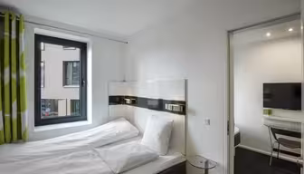 Lyst, moderne hotelværelse med dobbeltseng, hvidt sengetøj og vindue med grønne og hvide gardiner. Tilstødende værelse synligt gennem en åben dør, der viser et tv og et skrivebord med en stol. Væggene er hvide, hvilket forstærker rummets minimalistiske indretning.