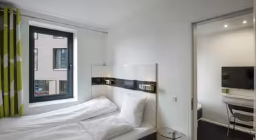 Lyst, moderne hotelværelse med dobbeltseng, hvidt sengetøj og vindue med grønne og hvide gardiner. Tilstødende værelse synligt gennem en åben dør, der viser et tv og et skrivebord med en stol. Væggene er hvide, hvilket forstærker rummets minimalistiske indretning.