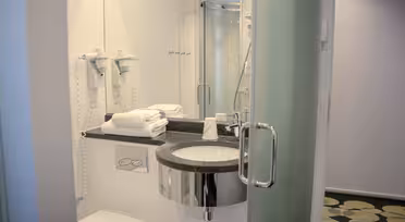 Et moderne badeværelse med en væghængt vask, et rundt spejl og en bruseniche i glas. To hårtørrere er på væggen, og håndklæder er pænt stablet på disken. Gulvet har et geometrisk mønster, og en grøn glasdør er delvist åben.
