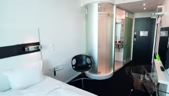 Moderne hotelværelse med en pænt redt seng, bruser i klart glas og en minimalistisk sort stol. Et tv er monteret på væggen overfor sengen, og et glasbord er i nærheden. Rummet har et slankt, moderne design med hvide og sorte accenter.