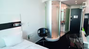 Moderne hotelværelse med en pænt redt seng, bruser i klart glas og en minimalistisk sort stol. Et tv er monteret på væggen overfor sengen, og et glasbord er i nærheden. Rummet har et slankt, moderne design med hvide og sorte accenter.