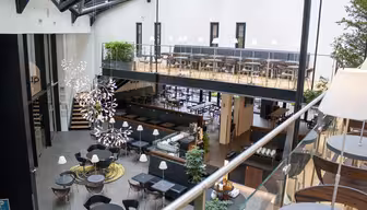Moderne cafe med to etager med moderne møbler. Det nederste niveau har runde borde og stole, mens det øverste niveau har båse med hynder. Store vinduer og metalbjælker giver en åben, luftig atmosfære. Dekorativ belysning og planter forbedrer rummet.