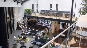 Moderne cafe med to etager med moderne møbler. Det nederste niveau har runde borde og stole, mens det øverste niveau har båse med hynder. Store vinduer og metalbjælker giver en åben, luftig atmosfære. Dekorativ belysning og planter forbedrer rummet.