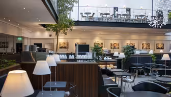 Moderne, stilfuld cafe med slanke sorte borde og stole, hvide lamper og grønne indendørs planter. Ovenpå pryder flere siddepladser og kunst på væggen. Store vinduer oplyser rummet med naturligt lys, hvilket skaber en indbydende atmosfære.