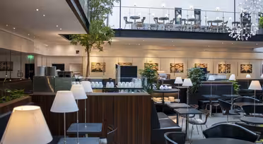 Moderne, stilfuld cafe med slanke sorte borde og stole, hvide lamper og grønne indendørs planter. Ovenpå pryder flere siddepladser og kunst på væggen. Store vinduer oplyser rummet med naturligt lys, hvilket skaber en indbydende atmosfære.