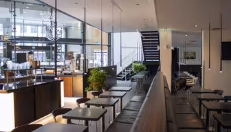 Moderne café interiør med slanke sorte borde og stole. En trappe fører til et øverste niveau. Store vinduespartier slipper naturligt lys ind, og pendullamper hænger fra loftet. Grønne planter tilføjer et strejf af natur til det minimalistiske design.