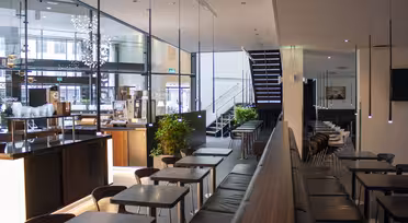 Moderne café interiør med slanke sorte borde og stole. En trappe fører til et øverste niveau. Store vinduespartier slipper naturligt lys ind, og pendullamper hænger fra loftet. Grønne planter tilføjer et strejf af natur til det minimalistiske design.