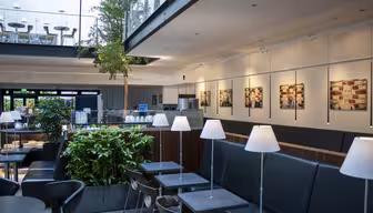 Moderne cafeinteriør med slanke sorte siddepladser og borde. Hvide pendler hænger over hovedet. Grønt og indrammede billeder pryder rummet, mens en glasbalkon med ekstra siddepladser har udsigt over området. Naturligt lys filtrerer ind fra vinduerne.