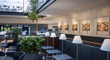 Moderne cafeinteriør med slanke sorte siddepladser og borde. Hvide pendler hænger over hovedet. Grønt og indrammede billeder pryder rummet, mens en glasbalkon med ekstra siddepladser har udsigt over området. Naturligt lys filtrerer ind fra vinduerne.