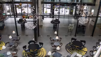 Et moderne, rummeligt caféinteriør med runde borde, sorte stole og abstrakte gule tæpper. Store glasruder giver udsigt til parkerede biler udenfor. Unikke lysekroner hænger fra loftet og tilføjer elegance til rummet.