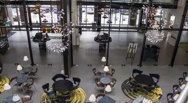 Et moderne, rummeligt caféinteriør med runde borde, sorte stole og abstrakte gule tæpper. Store glasruder giver udsigt til parkerede biler udenfor. Unikke lysekroner hænger fra loftet og tilføjer elegance til rummet.