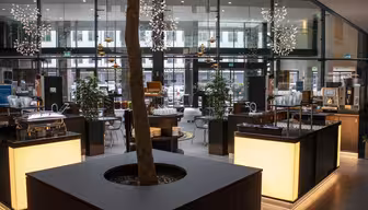 Moderne cafe med slanke bordplader, en central træplantemaskine og pendellamper. Glasvægge giver udsigt til bymæssige omgivelser. Forskellige siddemuligheder og selvbetjeningsstationer er synlige, hvilket skaber en afslappet atmosfære.