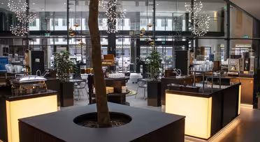 Moderne cafe med slanke bordplader, en central træplantemaskine og pendellamper. Glasvægge giver udsigt til bymæssige omgivelser. Forskellige siddemuligheder og selvbetjeningsstationer er synlige, hvilket skaber en afslappet atmosfære.
