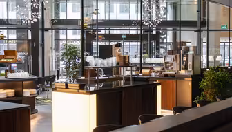Moderne cafeinteriør med store glasvinduer, slanke træborde, pendellamper og forskellige planter. Stabler af hvide tallerkener og en kaffemaskine står på disken. Atmosfæren er lys og indbydende.