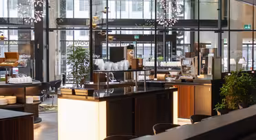 Moderne cafeinteriør med store glasvinduer, slanke træborde, pendellamper og forskellige planter. Stabler af hvide tallerkener og en kaffemaskine står på disken. Atmosfæren er lys og indbydende.