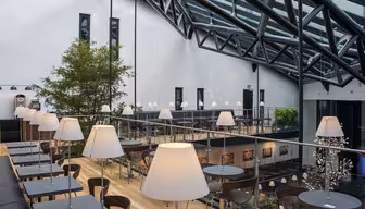 En moderne, stilfuld café med højt glasloft, sorte borde og hvide lamper. Der er flere siddepladser på forskellige niveauer. Et stort træ og kunst tilføjer til indretningen. Naturligt lys oversvømmer rummet fra loftsvinduerne.