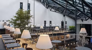 En moderne, stilfuld café med højt glasloft, sorte borde og hvide lamper. Der er flere siddepladser på forskellige niveauer. Et stort træ og kunst tilføjer til indretningen. Naturligt lys oversvømmer rummet fra loftsvinduerne.