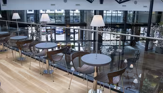 Moderne cafeinteriør på mezzaninplan med runde borde og træstole. Gulv til loft-vinduer har udsigt over en bygade. Blød belysning og dekorative planter skaber en hyggelig stemning.