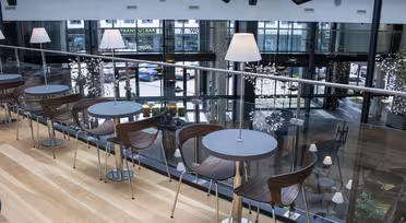 Moderne cafeinteriør på mezzaninplan med runde borde og træstole. Gulv til loft-vinduer har udsigt over en bygade. Blød belysning og dekorative planter skaber en hyggelig stemning.