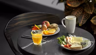 Et lille rundt bord med et morgenmadspålæg: et glas appelsinjuice, et hvidt krus kaffe, en tallerken med forskellige skåret frugter og en anden tallerken med ost, brød, salat og snittede grøntsager. En farverig plante er i baggrunden.