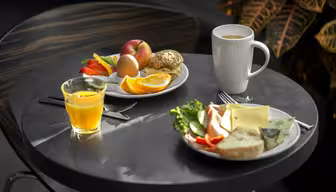 Et bord med to tallerkener med morgenmadsprodukter. En tallerken har en rulle, et æg, æbleskiver, appelsinskiver og rød peber. Den anden har en rulle med ost, salat og agurk. Der er et krus kaffe og et glas appelsinjuice.