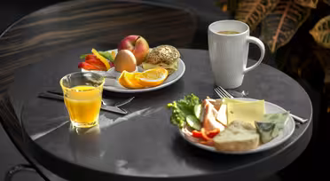 Et bord med to tallerkener med morgenmadsprodukter. En tallerken har en rulle, et æg, æbleskiver, appelsinskiver og rød peber. Den anden har en rulle med ost, salat og agurk. Der er et krus kaffe og et glas appelsinjuice.