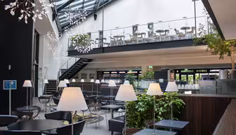 Moderne atrium med siddepladser med slanke sorte borde og stole på begge etager. Moderne hvide lamper og hængende lysarmaturer dekorerer rummet. Grønne planter tilføjer et strejf af natur, og en trappe forbinder de to etager.
