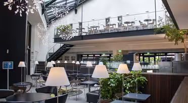Moderne atrium med siddepladser med slanke sorte borde og stole på begge etager. Moderne hvide lamper og hængende lysarmaturer dekorerer rummet. Grønne planter tilføjer et strejf af natur, og en trappe forbinder de to etager.